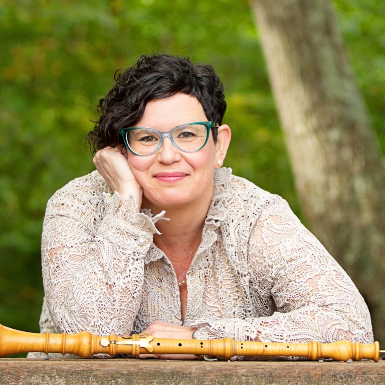 Woman in a white shirt resting her head on her hand with an oboe resting in front of her and outdoor greenery in the background.