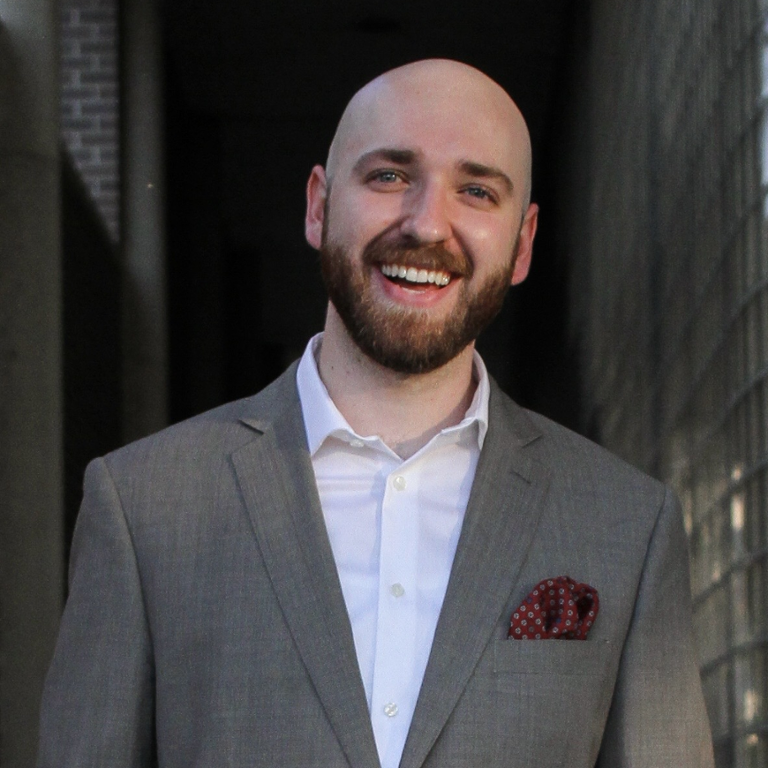 Waist-up, outdoor shot of a bearded man smiling, wearing a white shirt and gray suit jacket.